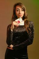 brunette fille dans noir robe montrant deux rouge frites et as, posant contre coloré studio Contexte. jeu, poker, casino. fermer. photo