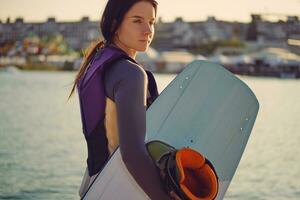 brunette fille dans une gris col roulé, spécial des sports gilet et bleu denim short est posant avec sa wakeboard sur une jetée de le côtier zone. fermer. photo