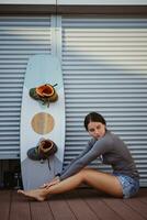 brunette fille dans gris col roulé et denim short est posant avec sa wakeboard séance sur sol à une côtier zone. sport et des loisirs. photo