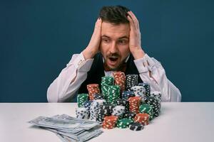 homme dans noir gilet et chemise séance à blanc table avec piles de frites et en espèces sur il, posant sur bleu Contexte. jeu, poker, casino. fermer. photo