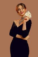brunette femme, nu épaules, dans noir robe et bijoux. souriant, montrant deux en jouant cartes, posant sur marron Contexte. poker, casino. fermer photo