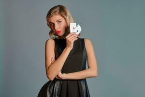blond fille dans noir élégant robe montrant deux en jouant cartes, posant contre gris Contexte. jeux d'argent divertissement, poker, casino. fermer. photo