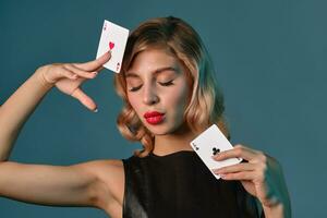 blond fille dans noir cuir robe montrant deux en jouant cartes, posant contre bleu Contexte. jeux d'argent divertissement, poker, casino. fermer. photo