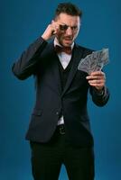 homme dans noir classique costume et rouge nœud papillon montrant un puce et certains dollar factures, posant sur bleu Contexte. jeu, poker, casino. fermer. photo