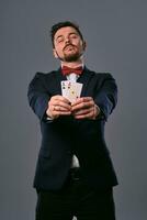 homme dans noir classique costume et rouge nœud papillon montrant deux en jouant cartes tandis que posant contre gris studio Contexte. jeu, poker, casino. fermer. photo