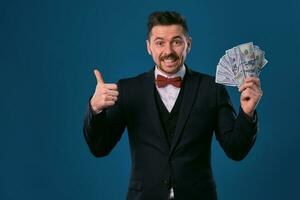 homme dans noir costume et rouge nœud papillon est montrant une ventilateur de cent dollar factures, posant sur bleu studio Contexte. jeu, poker, casino. fermer. photo