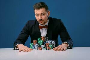 homme dans noir costume est séance à blanc table avec coloré piles de frites sur il, posant sur bleu studio Contexte. jeu, poker, casino. fermer. photo