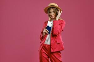 blond fille dans paille chapeau, blanc chemisier et rouge tailleur-pantalon. elle est en portant passeport et billet tandis que posant contre rose studio Contexte. fermer photo