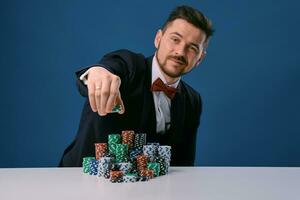 homme dans noir costume est séance à blanc table avec coloré piles de frites sur il, posant sur bleu studio Contexte. jeu, poker, casino. fermer. photo