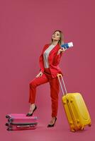 blond femme dans rouge tailleur-pantalon, blanc chemisier, noir talons. elle mettre sa jambe sur valise, Jaune un proche, en portant passeport et billet, rose Contexte photo