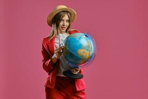 blond Dame dans paille chapeau, blanc chemisier et rouge tailleur-pantalon. elle est en portant une globe, passeport et billet tandis que posant contre rose Contexte. fermer photo