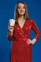 blond femme avec se maquiller, dans rouge paillette robe est souriant, montrant deux as, posant sur bleu Contexte. jeu, poker, casino. fermer. photo