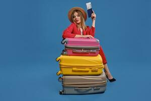 blond femme dans paille chapeau, blanc chemisier, rouge tailleur-pantalon. séance penché sur coloré les valises, en portant passeport et billet, posant sur bleu Contexte photo