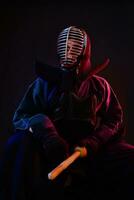 proche en haut. kendo combattant portant dans un armure, traditionnel kimono, casque, séance, pratiquant martial art avec shinaï bambou épée, noir Contexte. photo