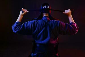 proche en haut tir, kendo combattant portant dans un armure et traditionnel kimono est attacher le laçage sur le sien casque permanent retour contre une noir Contexte. photo