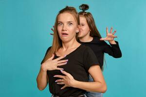 maman et fille avec une marrant coiffures, habillé dans noir chemises et bleu denim jeans sont posant contre une bleu studio Contexte. fermer tir. photo