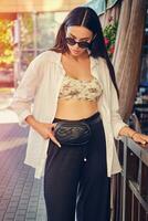 portrait de une fille dans foncé des lunettes de soleil posant dans ville près été terrasse. habillé dans Haut avec floral imprimer, blanc chemise, noir pantalon, taille sac. photo