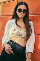 portrait de fille dans foncé des lunettes de soleil posant dans ville contre Orange bâtiment. habillé dans Haut avec floral imprimer, blanc chemise, noir pantalon, taille sac. photo