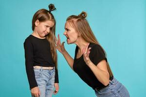 maman et fille avec une marrant coiffures, habillé dans noir chemises et bleu denim jeans sont posant contre une bleu studio Contexte. fermer tir. photo