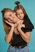 maman et fille avec une marrant coiffures, habillé dans noir chemises et bleu denim jeans sont posant contre une bleu studio Contexte. fermer tir. photo