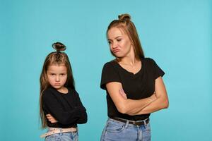 maman et fille avec une marrant coiffures, habillé dans noir chemises et bleu denim jeans sont posant contre une bleu studio Contexte. fermer tir. photo