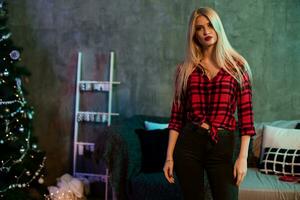 portrait de une magnifique blond femme dans une Père Noël chapeau, rouge chemise dans une cage et noir jeans à Noël fête à maison. photo