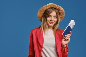 blond fille dans paille chapeau, blanc chemisier, rouge tailleur-pantalon. elle est montrant passeport et billet, posant sur bleu Contexte. en voyageant concept. fermer photo