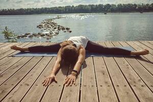 femme dans blanc maillot de corps et noir leggings est pratiquant yoga performant yoga-asanas sur une tapis en plein air, près une bord de rivière. photo