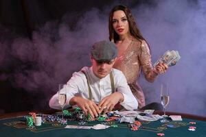 homme en jouant poker à casino séance à table avec piles de puces, argent, cartes. célébrer gagner avec mignonne femme. noir, fumée Contexte. fermer. photo