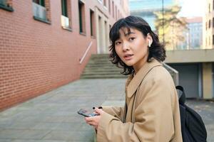 élégant moderne asiatique fille, étudiant dans sans fil écouteurs, des promenades autour ville avec sac à dos, souriant avec content visage photo