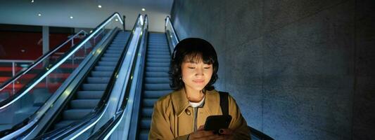 mignonne Jeune femme Aller vers le bas le escalier mécanique à le tube, en utilisant métro métro à commuer à travail ou université, permanent avec téléphone intelligent photo