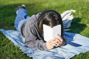 portrait de content fille en riant, couvrant visage avec sa livre, mensonge sur herbe dans parc photo