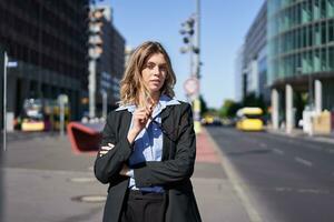 portrait de sur de soi affaires femme dans costume, traverser bras sur poitrine, à la recherche sûr de lui dans ville centre, permanent sur rue photo