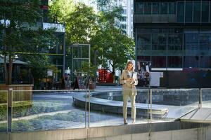 entreprise femme dans costume, permanent avec confiance dans beige costume avec travail les documents en plein air, posant dans ville centre photo