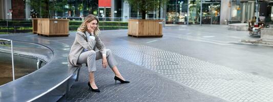 souriant femelle entrepreneur dans costume, Dame sur sa le déjeuner Pause attendre pour Quelqu'un près Fontaine sur vide rue photo