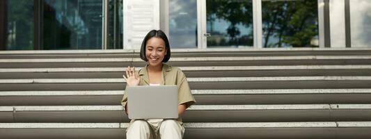 portrait de Jeune asiatique fille, étudiant pourparlers à ordinateur portable, vidéo discuter, Parlant pendant en ligne réunion, séance en plein air sur escaliers photo