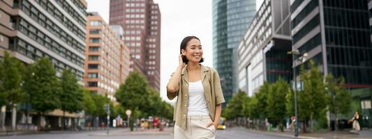 Extérieur coup de Jeune souriant asiatique femme, permanent sur rue dans jour, à la recherche autour, posant content photo