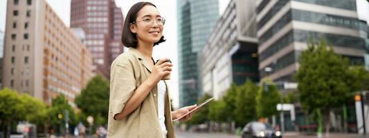 content femme explorant le ville. Jeune coréen fille détient tablette, les boissons café et des promenades le long de rue avec gros sourire sur sa visage photo