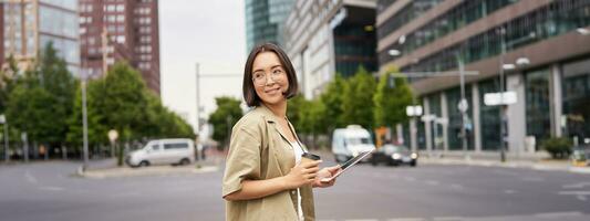 content femme explorant le ville. Jeune coréen fille détient tablette, les boissons café et des promenades le long de rue avec gros sourire sur sa visage photo