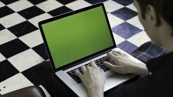 plus de le épaule vue de une homme en utilisant le sien ordinateur avec vert écran et séance sur une canapé, temps laps. Stock images. homme d'affaire dactylographie sur le sien portable avec chromakey sur à carreaux sol Contexte. photo