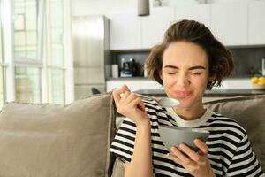 portrait de Jeune femelle modèle, étudiant en mangeant rapide petit-déjeuner, en portant bol de granola ou céréales avec lait, séance sur canapé dans vivant pièce photo