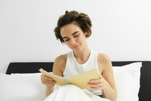 proche en haut portrait de Jeune femme, en train de lire journal, séance dans lit et en étudiant, réviser pour examen avant Aller à sommeil photo