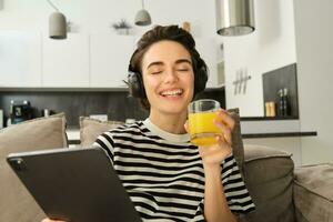 proche en haut portrait de Jeune brunette femme dans vivant chambre, en train de regarder marrant vidéo ou la télé séries sur numérique tablette, en buvant Orange jus, séance sur canapé et en riant photo