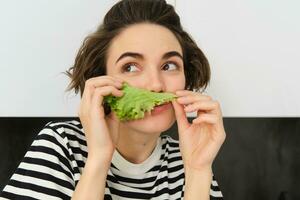 proche en haut de marrant mignonne femme, végétarien en mangeant salade feuille et souriant, concept de en bonne santé régime, fille aime légumes, permanent dans le cuisine photo