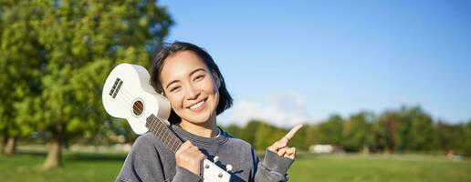 portrait de asiatique souriant fille, en portant ukulélé plus de épaule, montrer du doigt doigt à copie espace, bannière ou logo photo