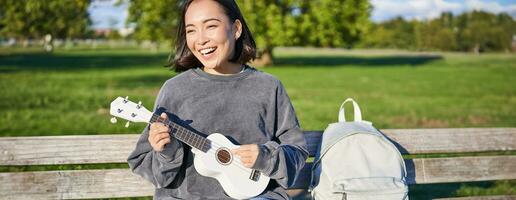 content mignonne fille est assis seul sur banc dans parc, pièces ukulélé guitare et jouit ensoleillé journée en plein air photo