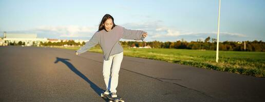 verticale portrait de content asiatique fille profiter planche à roulette amusement journée dehors. souriant coréen patineur sur longboard, équitation le long de vide rue sur ensoleillé journée photo