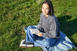 portrait de asiatique fille en train de lire livre, séance sur sa couverture dans parc, avec vert herbe, souriant Heureusement photo