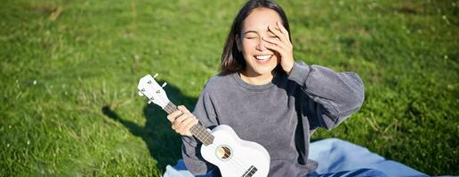 souriant asiatique fille avec ukulélé, en jouant dans parc et en chantant, mode de vie concept photo