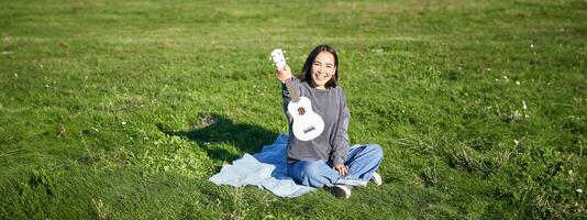 la musique et instruments. portrait de mignonne asiatique fille spectacles sa blanc ukulélé, pièces dans parc tandis que séance détendu sur couverture, profiter ensoleillé journée photo